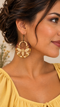 Cargar imagen en el visor de la galería, Model wearing Sunset Honey Bee lightweight wooden earrings with engraved bee and sunflower design, bronze-tone hooks, and a medium yellow top