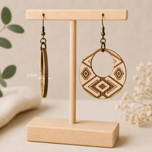 Cargar imagen en el visor de la galería, Side view of Tribal Spirit engraved wooden earrings on a light wood T-stand, showing the 2 mm American linden wood thickness beside a front-facing earring.