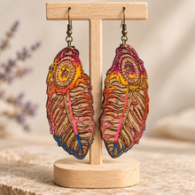 Cargar imagen en el visor de la galería, Boho feather wooden earrings displayed front-facing on a light wood stand, showing intricate laser engraving, hand-painted sunset colors and bronze-tone hooks.
