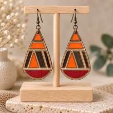Cargar imagen en el visor de la galería, Geometric teardrop hand-painted wooden earrings displayed front-facing on a light wood T-stand, showing orange, deep red, dark accents and bronze-tone hooks.
