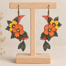 Cargar imagen en el visor de la galería, Moon Flower layered wooden earrings on a light wood stand with hand-painted orange and yellow flowers
