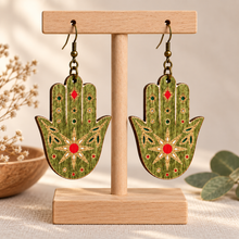 Cargar imagen en el visor de la galería, Mystical Hamsa Hand double-sided wooden earrings on a light wood stand with hand-painted green, red, gold, and white details
