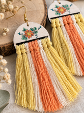Cargar imagen en el visor de la galería, Close-up of Sun Whisper wooden earrings showing the hand-painted floral design, gold-tone hooks, knotted macramé details and textured tassels

